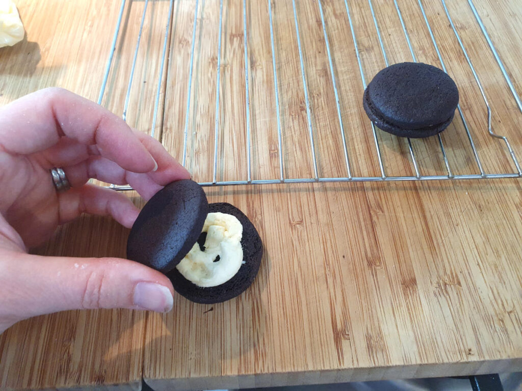 putting top cooking onto bottom cookie with vanilla filling pioped onto it, with write rack and completed fill cookie behind it.