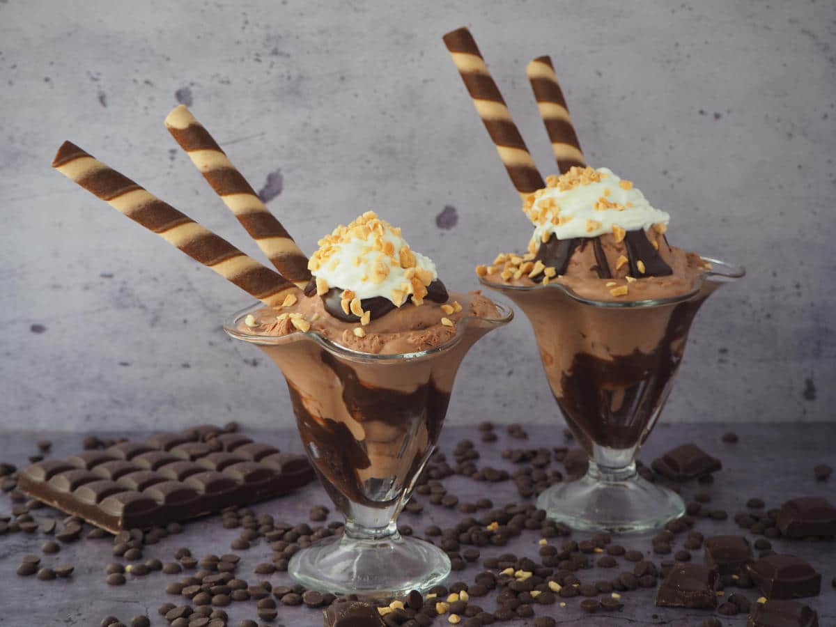 Side view of two chocolate sundaes in tall sundae glasses, with chocolate fudge sauce, chocolate ice cream, whipped cream, chopped nuts and chocolate and vanilla circular tube wafers, on a grey background, surrounded by chocolate pieces, chocolate chips and some chopped nuts.