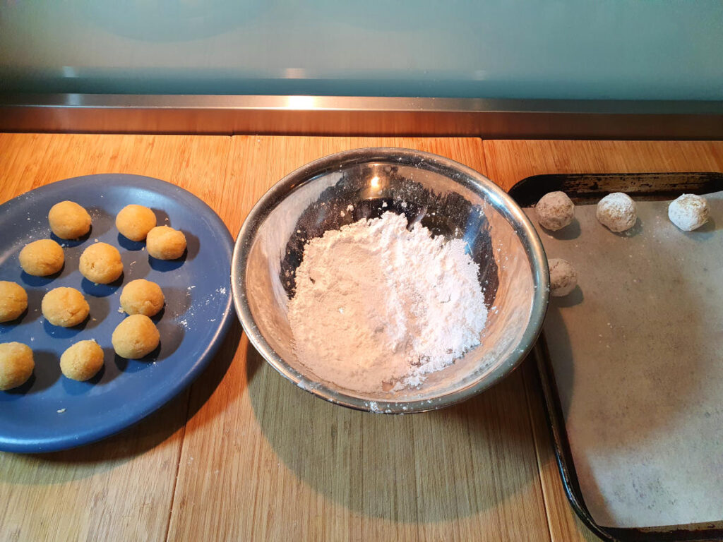 cookie production line showing rolled cookies on a plate on the left, bowl of sifted icing sugar in the middle and lined baking tray on the right.