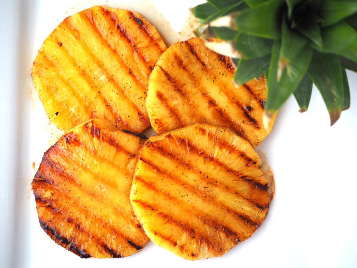 Top down view of four slices of grilled pineapple showing griddle lines, with a green pineapple top in the foreground out of focus, on a white plate.