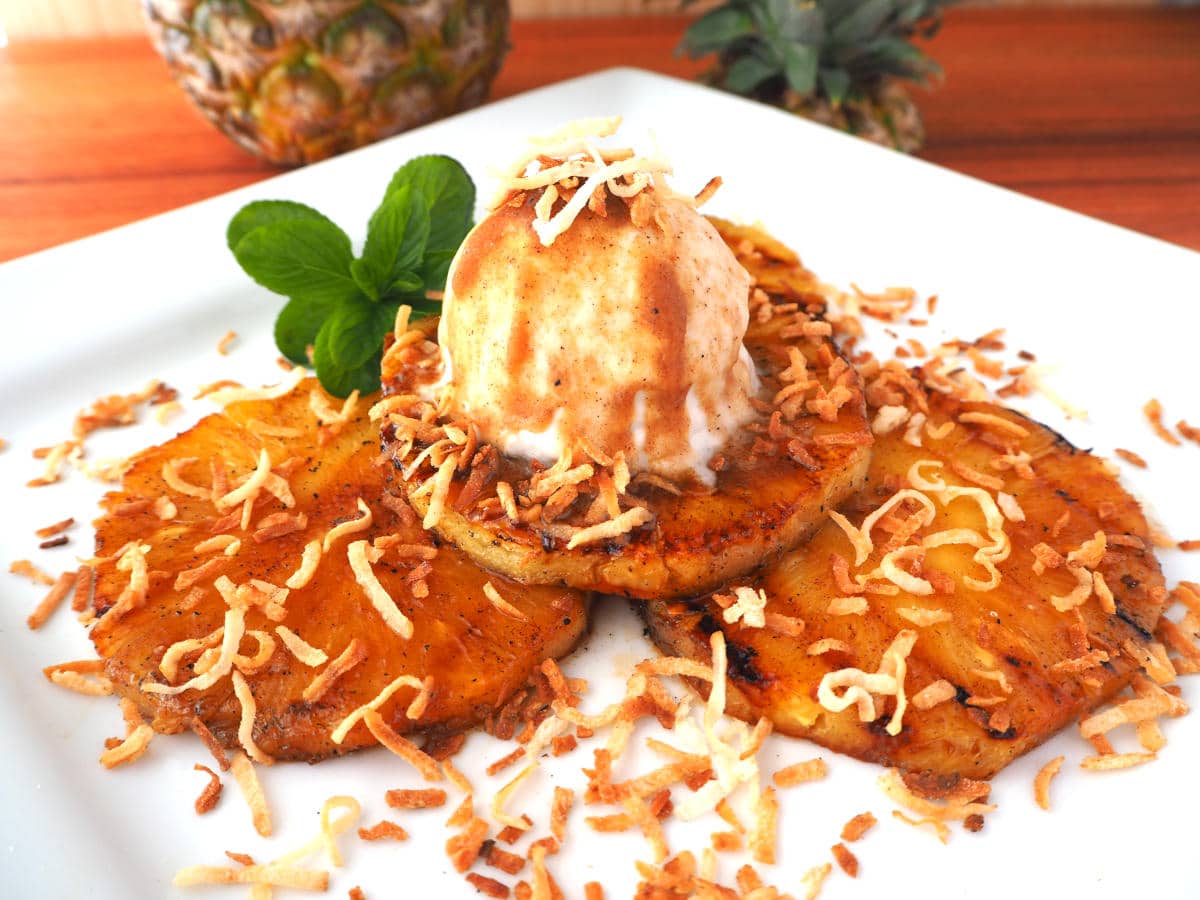 Side view of grilled pineapple with ice cream scoop and toasted shreaded coconut, with a mint sprig garnish, on a white plate, with a pineapple and pineapple top in the background.