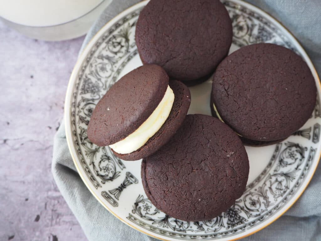 top down view of homemade oreas, with one on its side, on a white plate with a black border pattern and gold rim, on a grey tea towel and a glass of milk on the side.