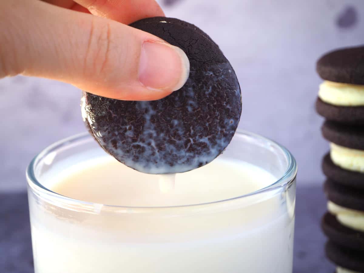 side view homemade oreo that's just been dunked in a glass of mil, with milk dripping off it and a stack of oreos to the side.