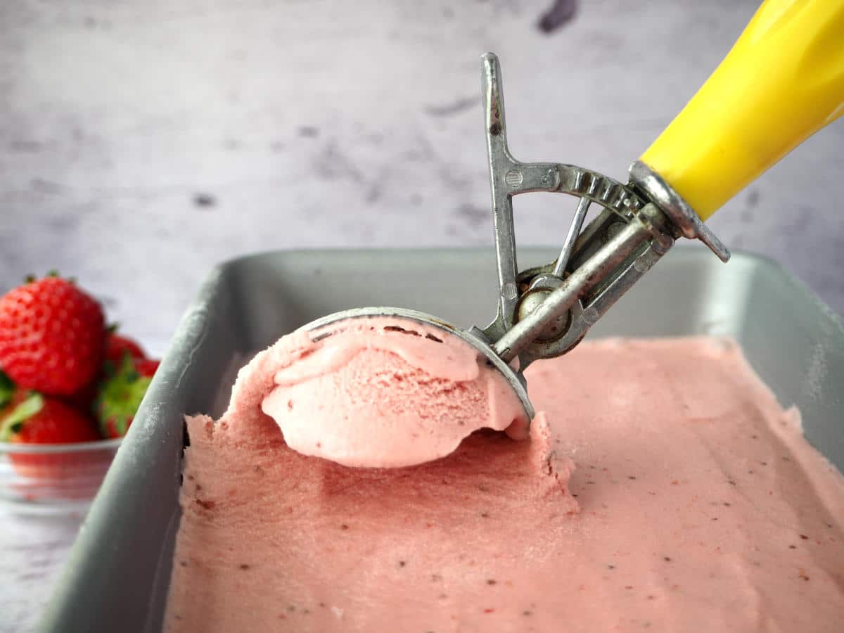 strawberry ice cream in pan being scooped with old fashioned metal and yellow plastic ice cream scoop, with bowl of strawberries in the background.