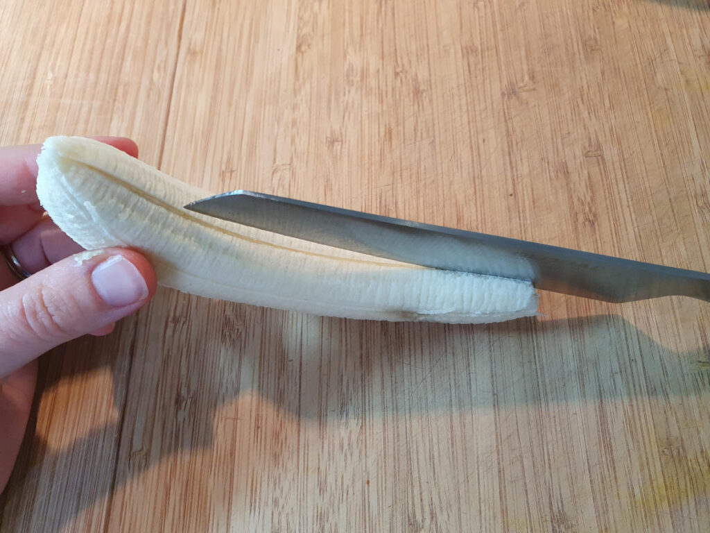 top down view spliting peeled banana lengthways in half with a sharp knif, on a wooded chopping board.