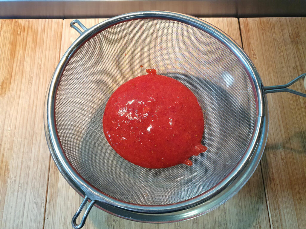 top down view showing blitzed stawberries being strained in a metal strainer over a glass mixing bowl.