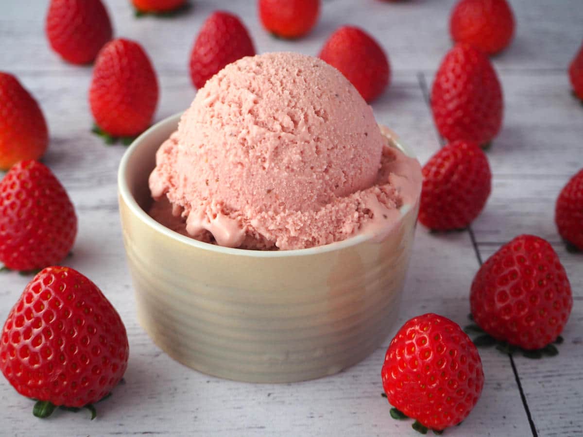 45 degree view of scoops of strawberry ice cream in a beige ramakin with strawberries scattered around it, on a white background.