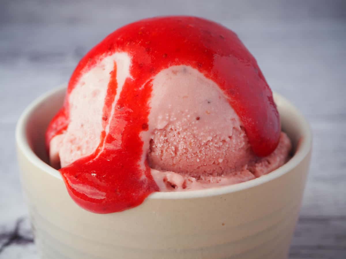 close up side view of scoop to slightly melted strawberry ice cream, with strawberry compote dripping over it and over the edge of the ramakin, on a white background.