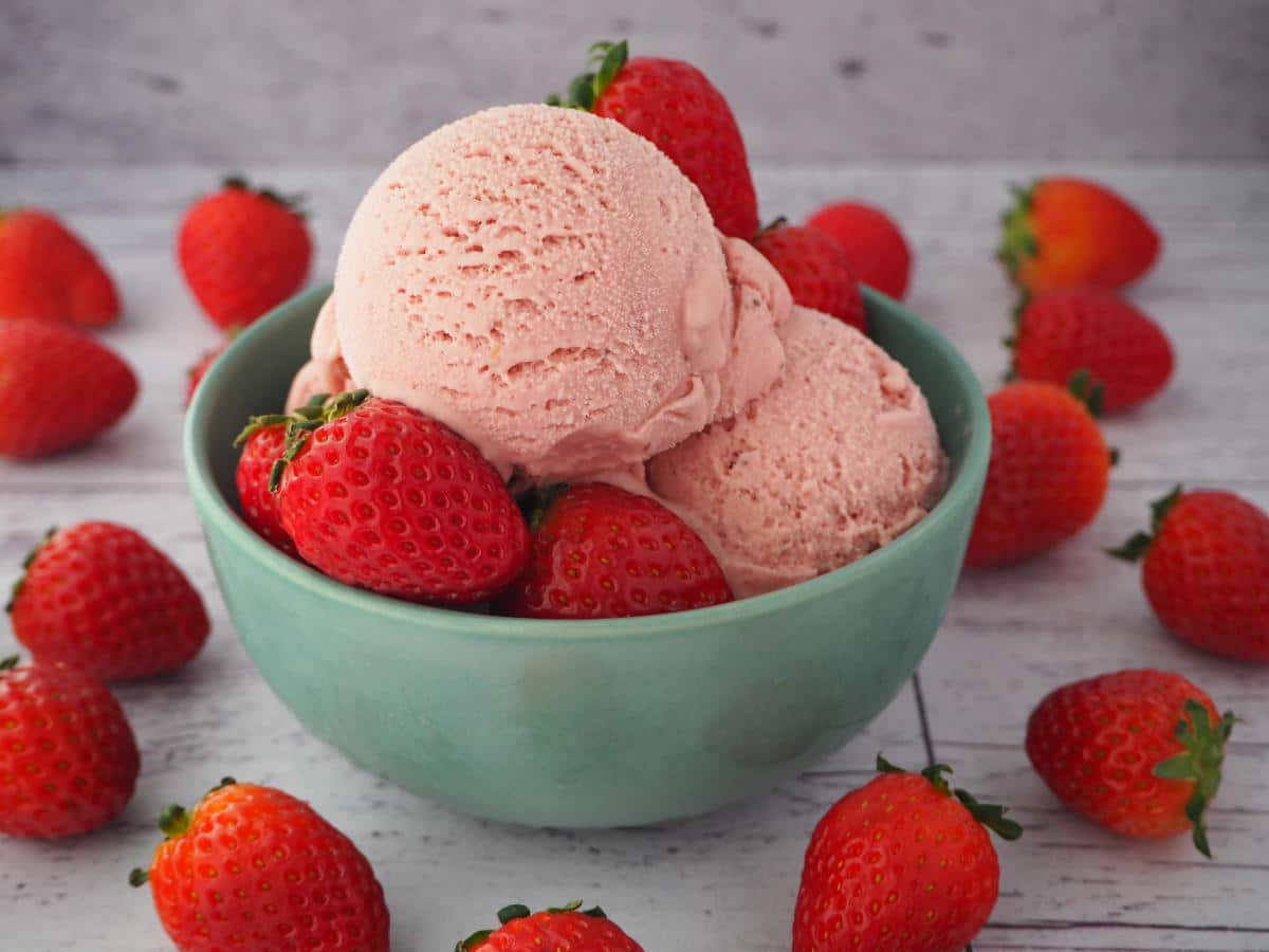 45 degree view of scoops of strawberry ice cream in a green bowl with strawberries in the bowl and strawberries scattered around it, on a white background.