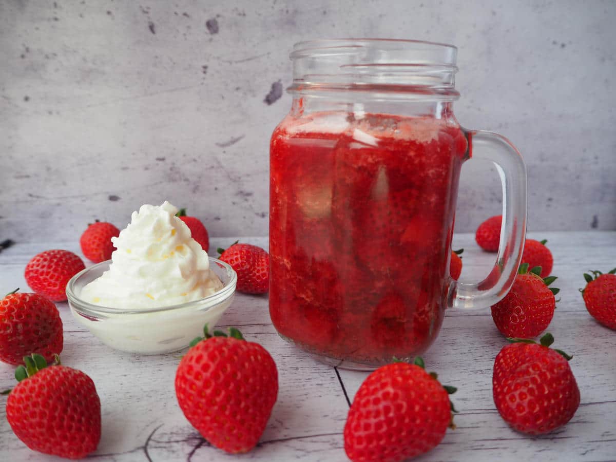 strawberry sauce in jar with whipped cream on side and strawberries scattered around, on a white floorboard surface.