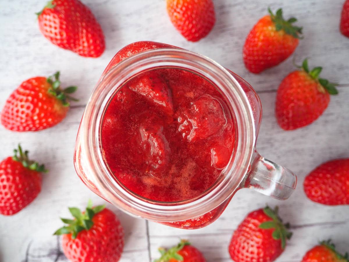 top down view of strawberry sauce in jar with strawberries scattered around, on a white floorboard surface.