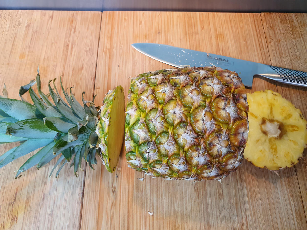 pineapple laid sidesways with the top and end cut off, with a large sharp knife, on a chopping board.