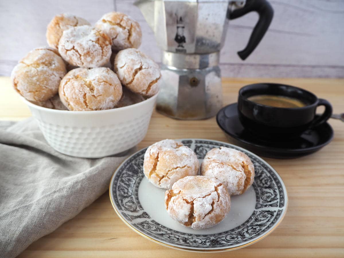 amaretti cookies on a plate, with bowl of amaretti cookies piled high, black epresso coffee cup with coffee and Italian stove top coffee pot in background.