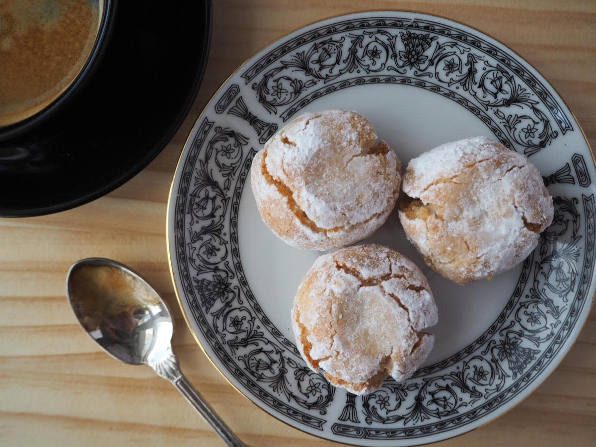 top down vew three amaretti cookies on a patterned plate, with cup of coffee and teaspoon on the side.