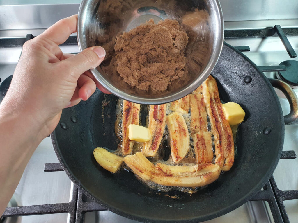 Adding brown sugar to bananas in frying pan on stove.