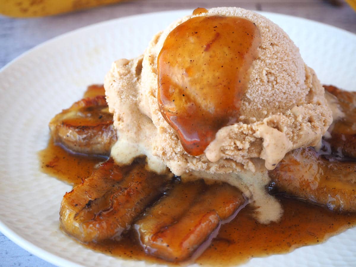 Caramelized bananas in caramel sauce, with a scoop of banana ice cream on top, and a spoon of caramel sauce dripping down the ice cream, on a white plate.