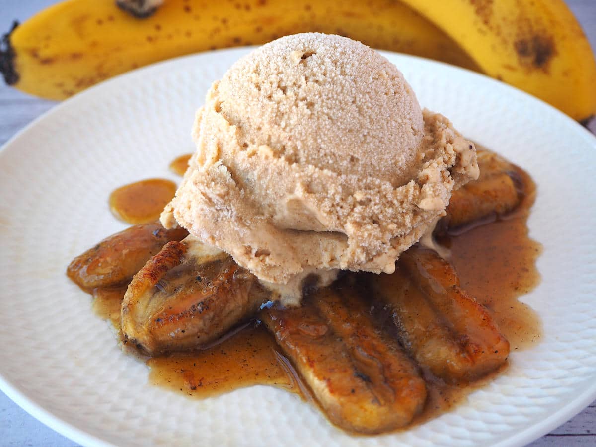 Caramelized bananas in caramel sauce, with a scoop of banana ice cream on top, on a white plate, with fresh bananas in the background.