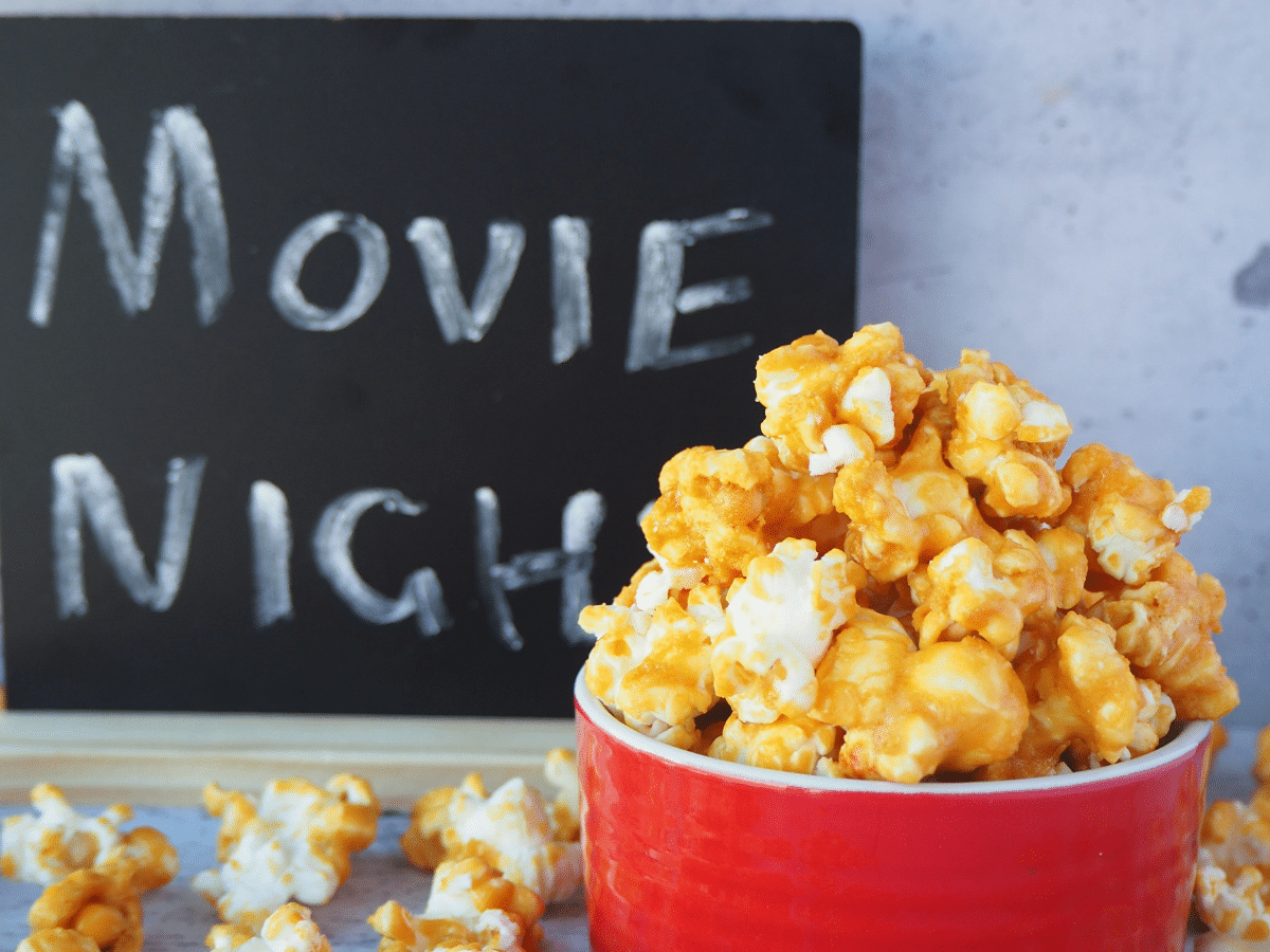 Caramel popcorn in a red bowl, with caramel popcorn scattered around it and a chalk board with Movie Night in the background.