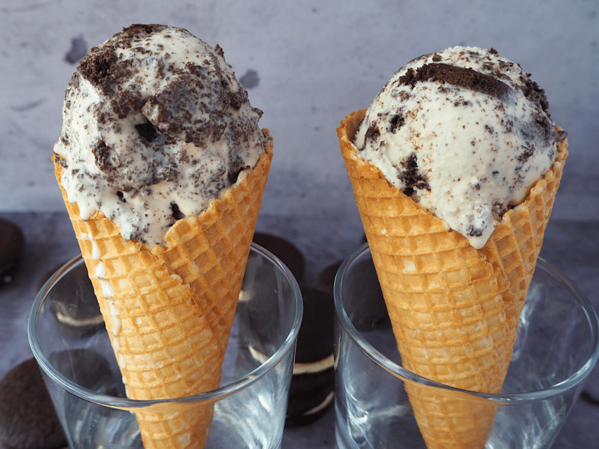 Two waffle ice cream cones with a large scoop of cookies and cream ice cream in each, resting in glasses, with Oreos scattered around them.