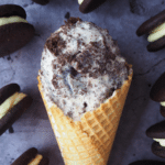 Close up of scoop of cookies and cream ice cream in a waffle cone, surrounded by Oreos.