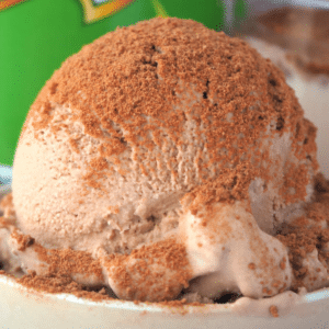 Close up scoop Milo ice cream sprinkled with Milo, in a small bowl, with Milo tin and glass of Milo in the background.