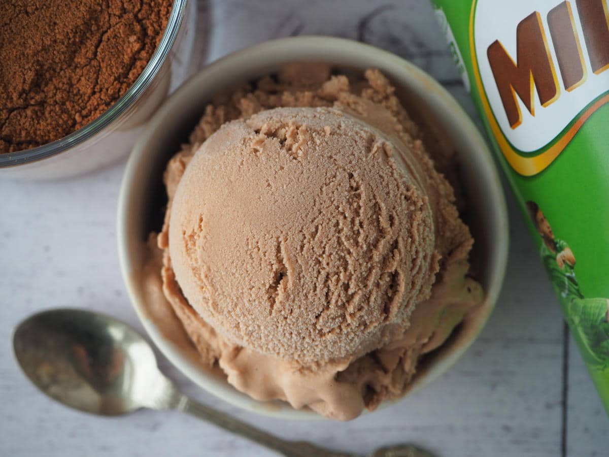 Top down view scoop of Milo ice cream in a bowl, with Milo tin on the side, glass of Milo and spoon.