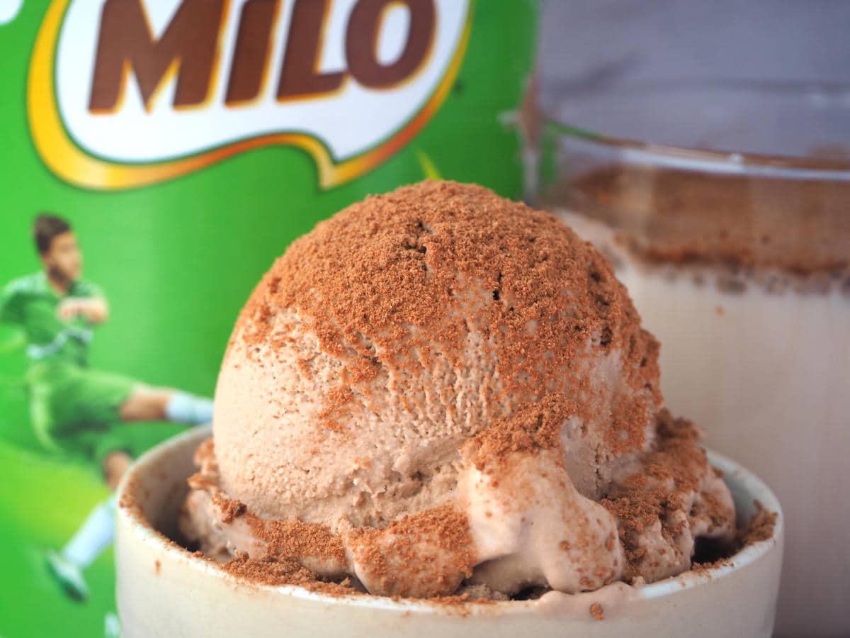 Close up scoop Milo ice cream sprinkled with Milo, in a small bowl, with Milo tin and glass of Milo in the background.