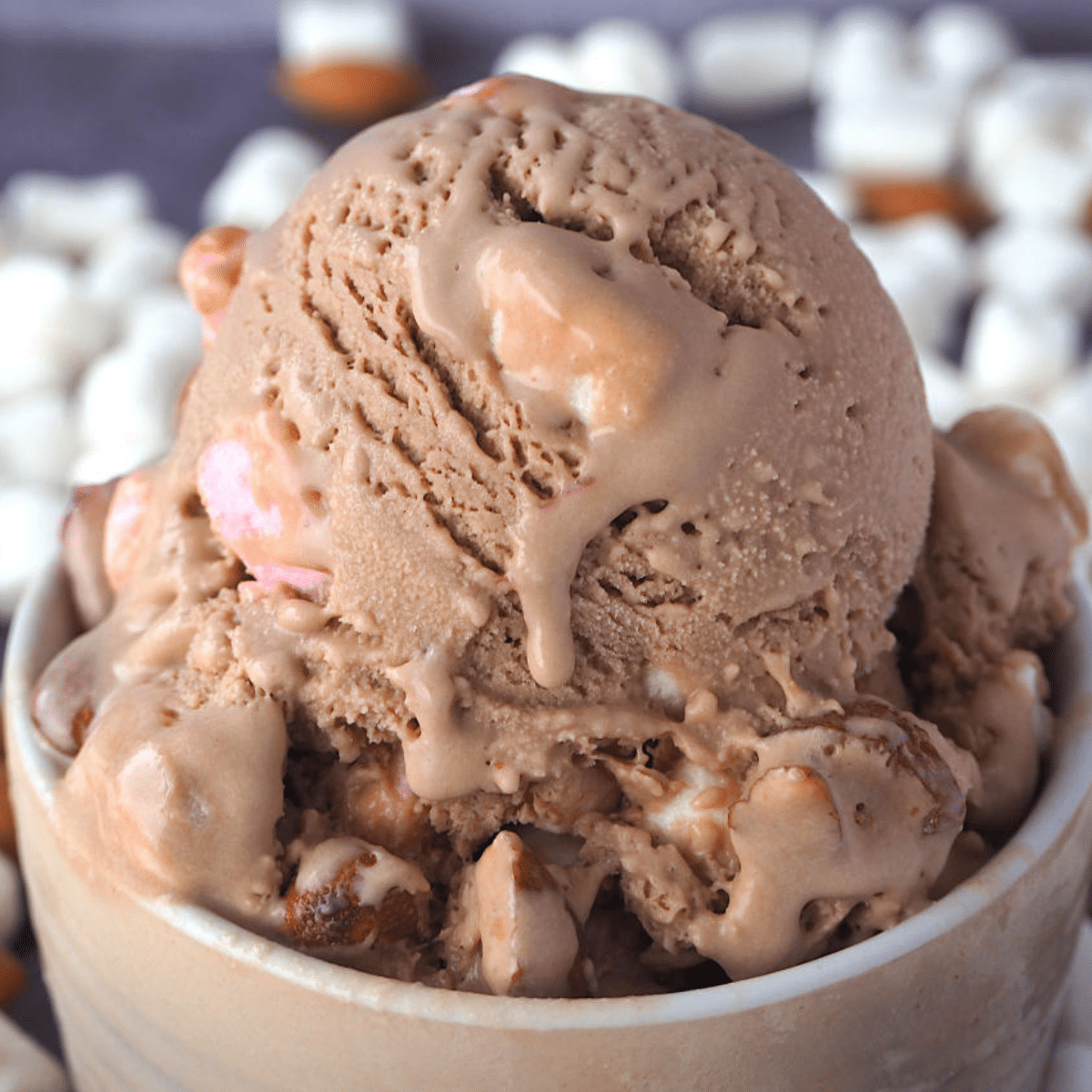 Close up scoop of rocky road ice cream with ice cream drip, with marshmallows and almonds in the background.