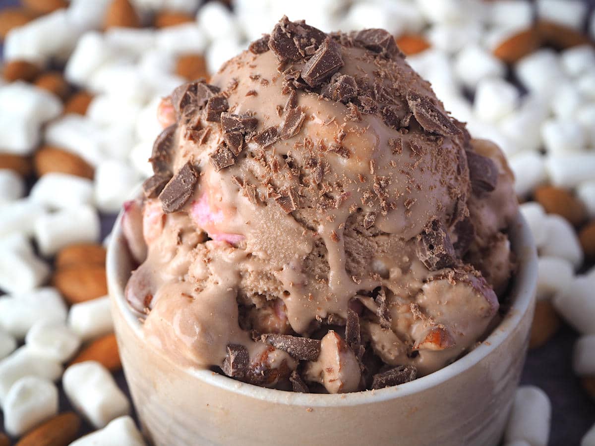 Close up scoop of rocky road ice cream with chopped chocolate sprinkled on top and ice cream drip, in a brown bowl, with marshmallows and almonds in the background.