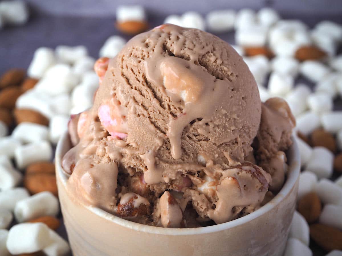 Close up scoop of rocky road ice cream with ice cream drip in a brown bowl, with marshmallows and almonds in the background.