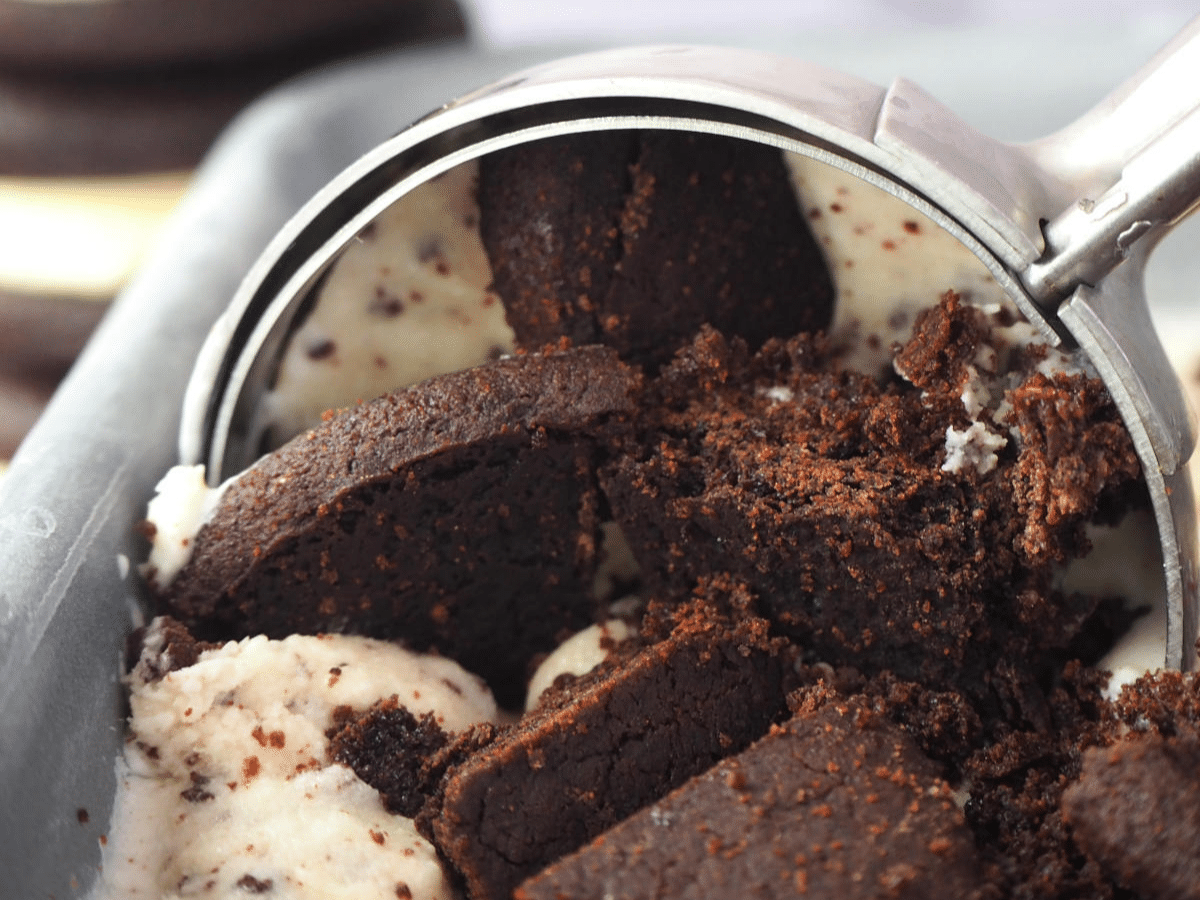 Close up scooping cookies and cream ice cream in loaf pan, with Oreos in the background.