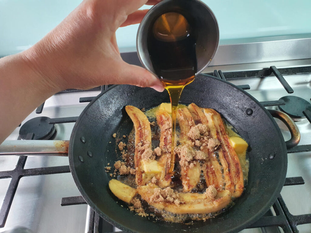 Adding golden syrup to bananas in frying pan on stove.