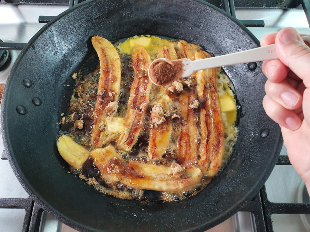 Adding ground nutmeg to bananas in frying pan on stove.