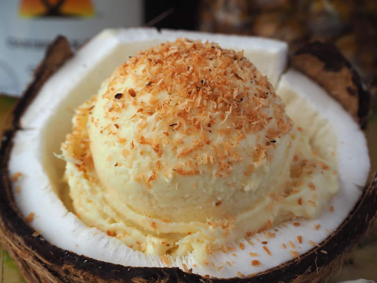 Scoop of pina cola ice cream sprinkled with toasted coconut in half a coconut, with pineapple and coconut rum bottle in background.