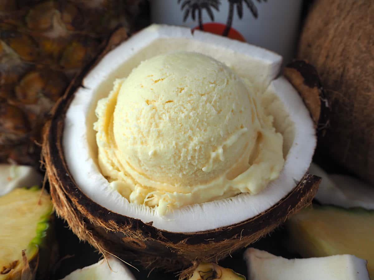 Scoop of pina cola ice cream in half a coconut, with pineapple and coconut rum bottle in background.