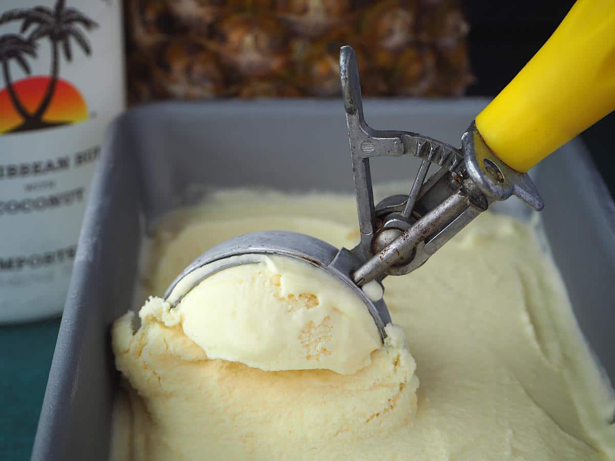 Scooping pina cola ice cream out of a pan with a metal ice cream scoop, with coconut rum and pineapple in the background.