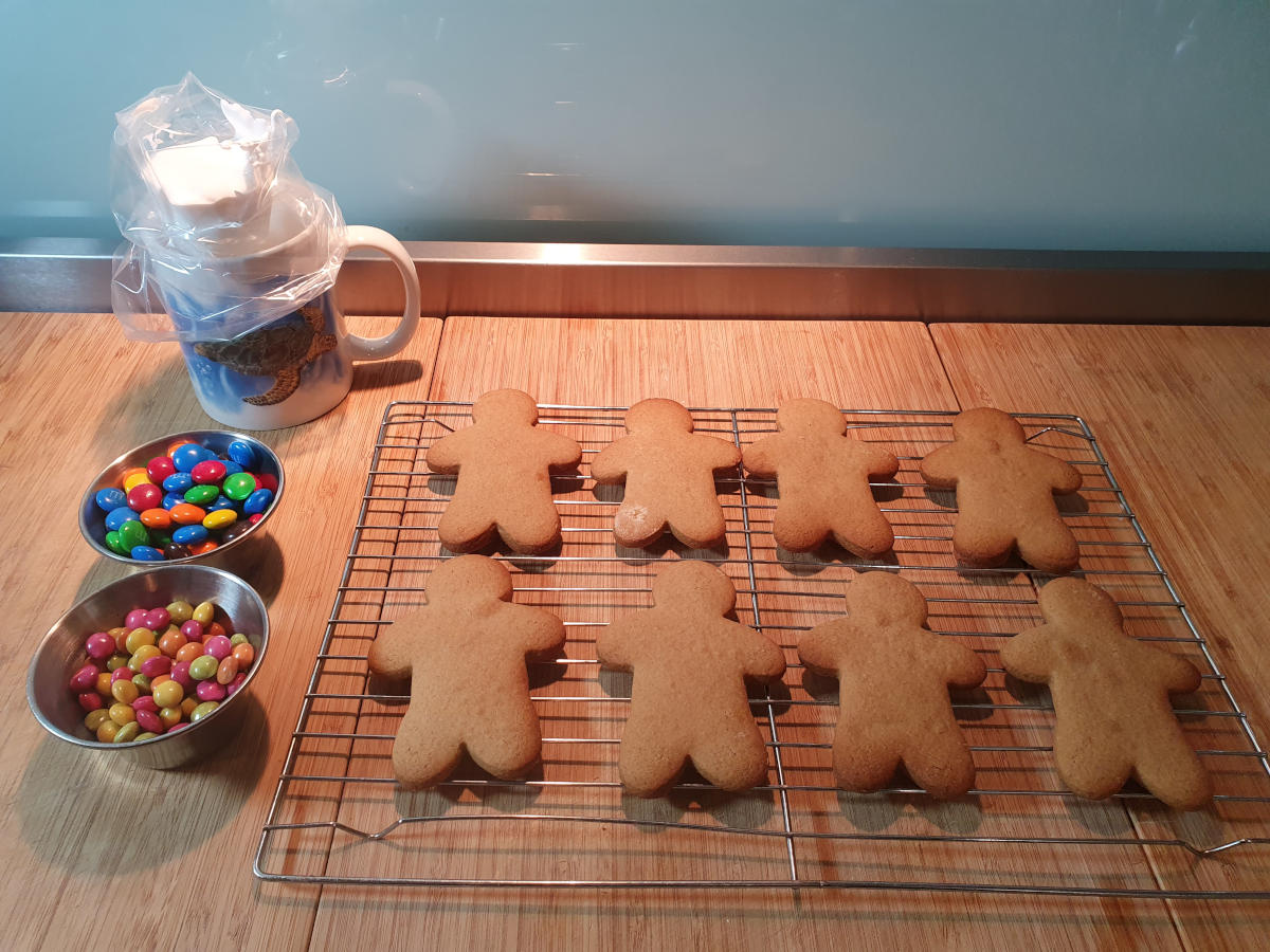 setting up to decorate gingerbread men, dishes if candies for buttons on the left, piping bag with icing mix in a mug to the left and wire rack of fully cooled gingerbread men ready to decorate.