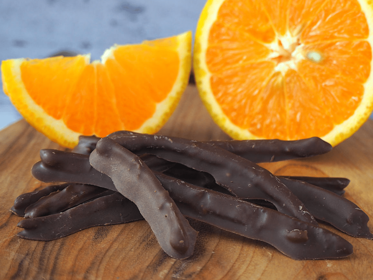 Chocolate covered orange peel with fresh slices of orange in background, on wooded chopping board.