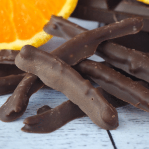 Close up of chocolate covered orange peel with slice of fresh orange and stacked dark chocolate in background.