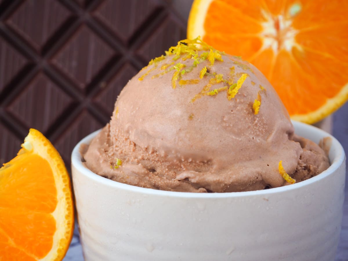 Scoop of chocolate orange ice cream in bowl, with orange zest on top and fresh orange slices in the front and background, and dark chocolate in the background.