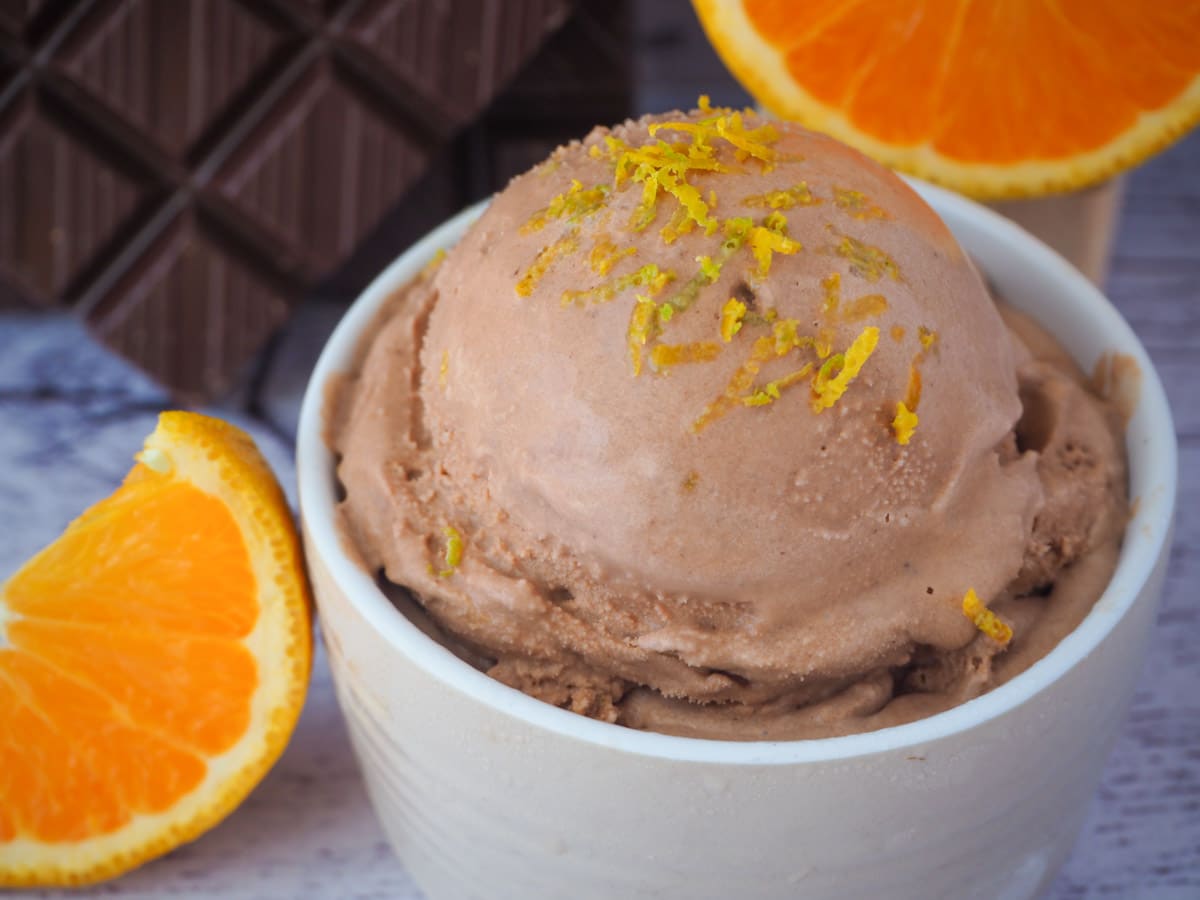 Scoop of slightly melting chocolate orange ice cream with orange zest on top, fresh orange slices and dark chocolate in the background.