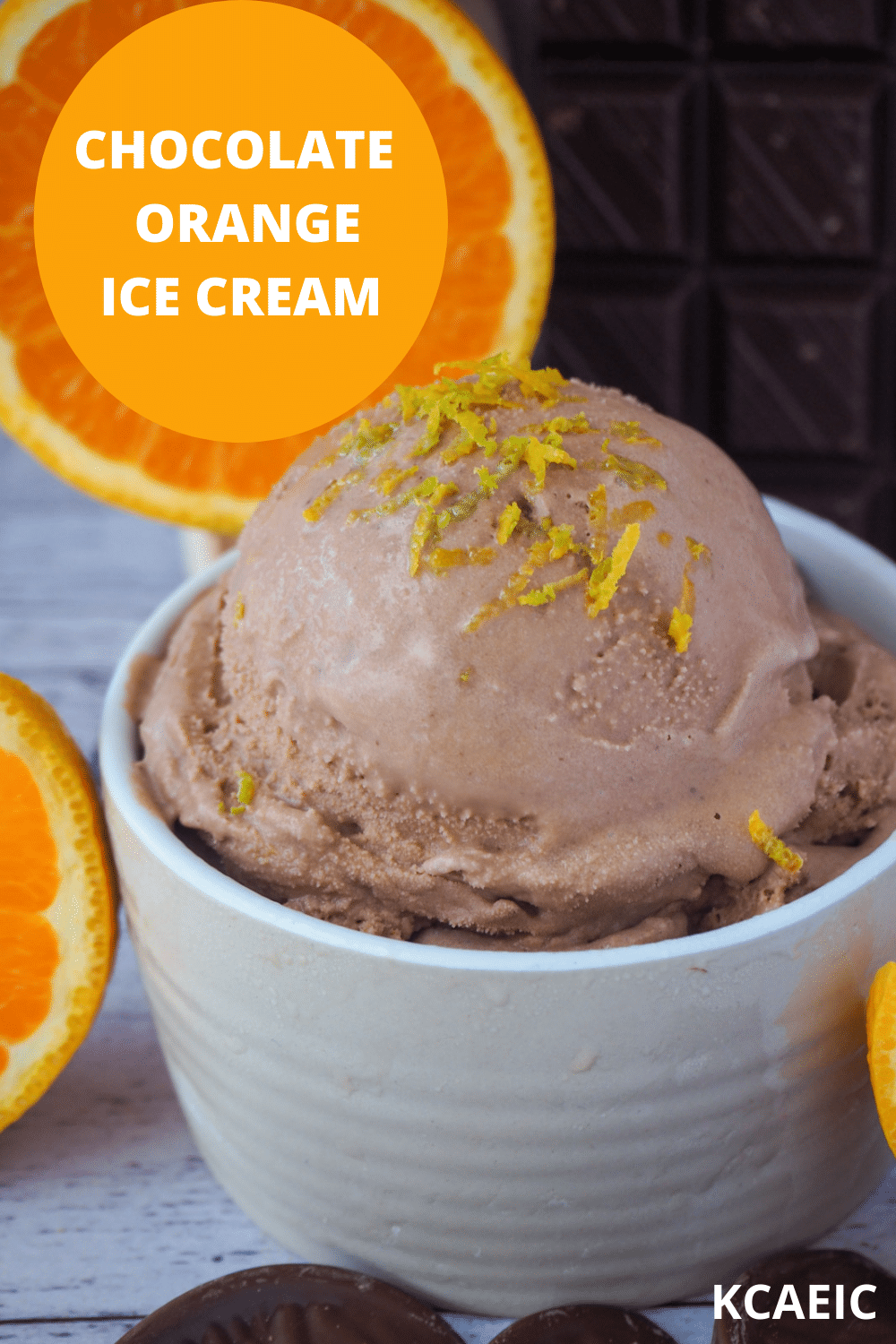 Scoop of chocolate orange ice cream in bowl, with orange zest on top and fresh orange and dark chocolate in the background, with text overlay, chocolate orange ice cream, KCAEIC.