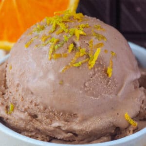 Close up scoop chocolate orange ice cream with orange zest on top, with fresh orange slice and dark chocolate in the background.