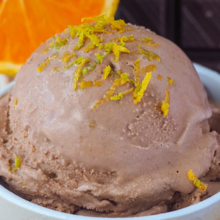 Close up scoop chocolate orange ice cream with orange zest on top, with fresh orange slice and dark chocolate in the background.