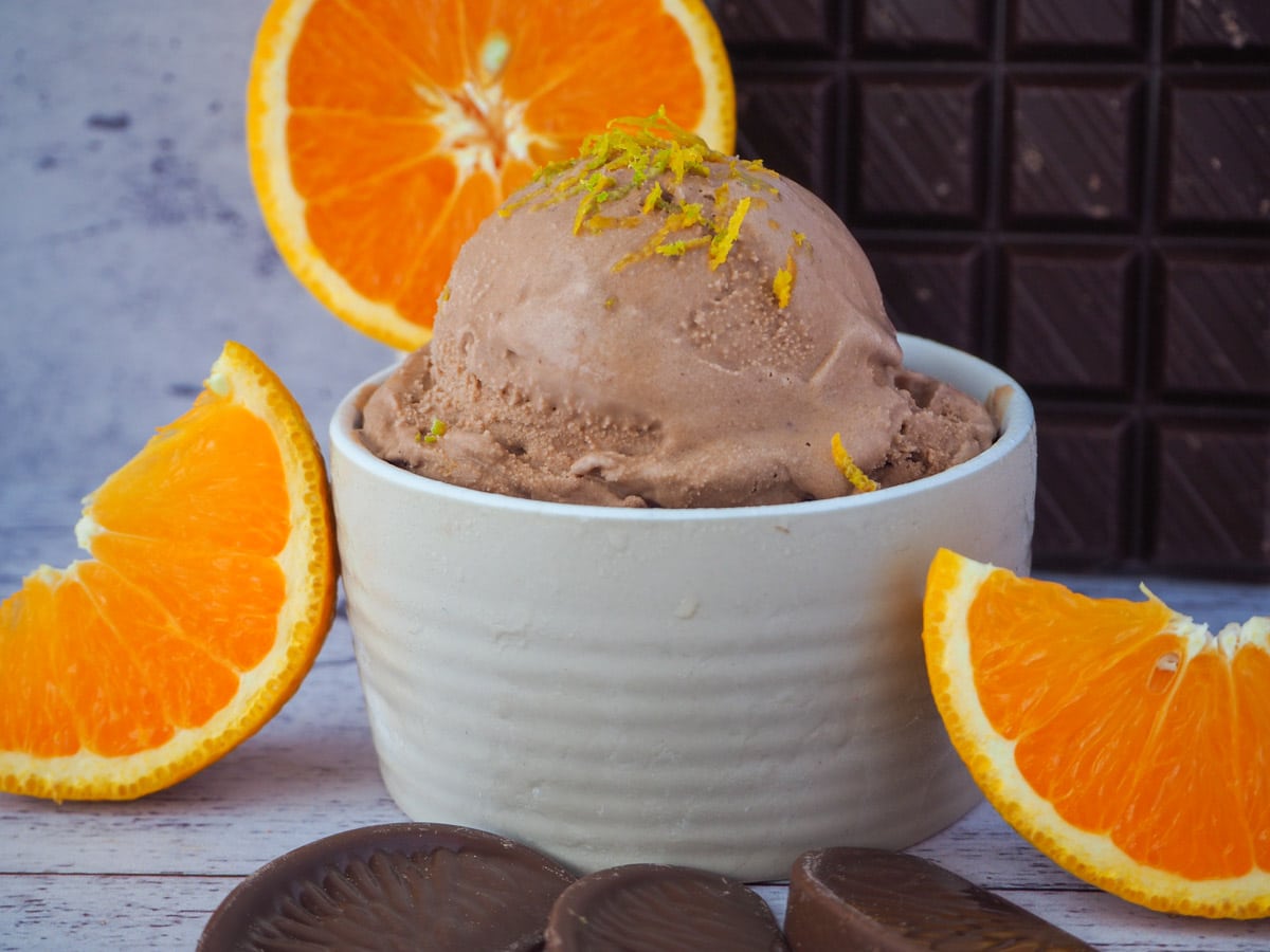 Scoop of chocolate orange ice cream in bowl, with orange zest on top and fresh orange slices in the front and background, and dark chocolate in the background.