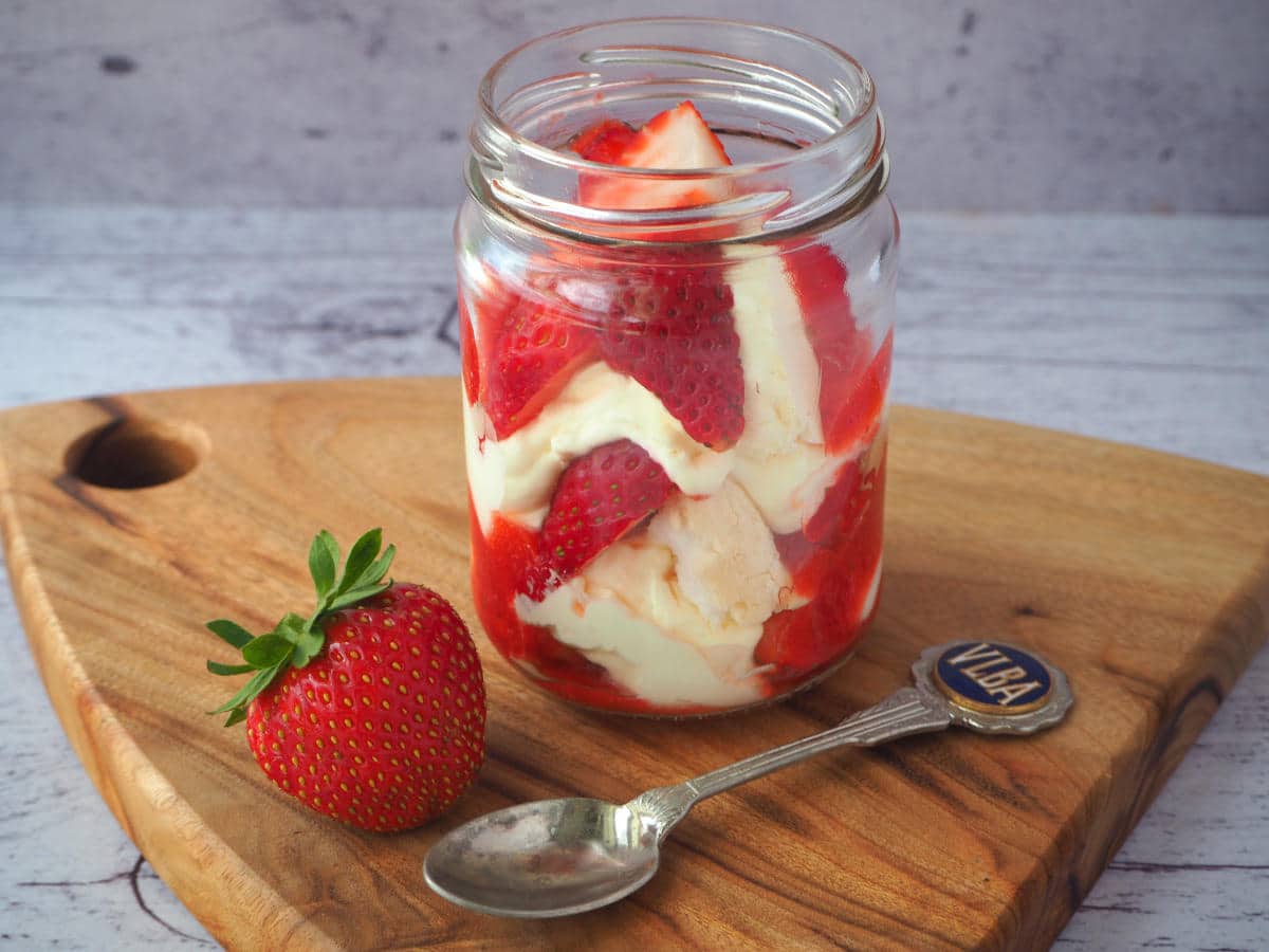 Jar of Eton mess on a board, with strawberry on the side and spoon.
