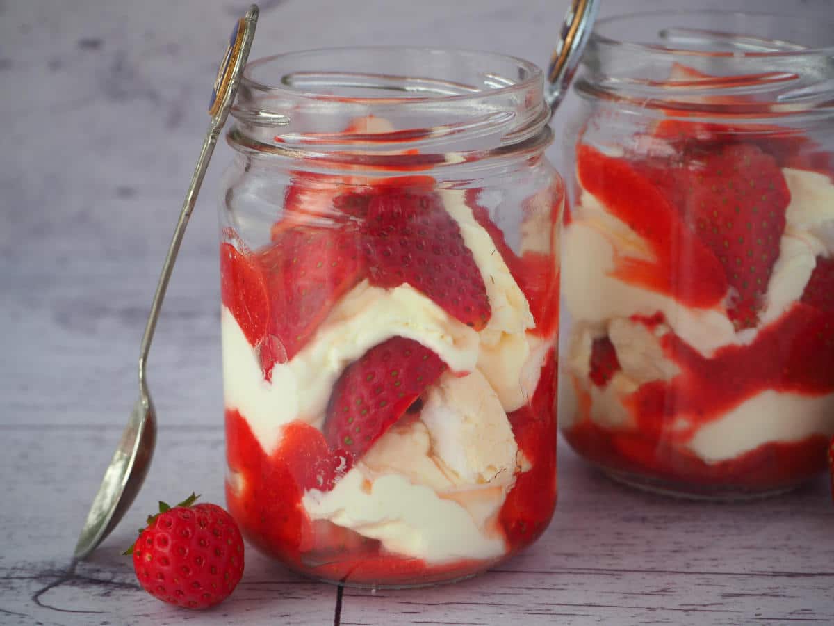 Two jars of Eton mess with fresh strawberries on the side and vintage spoons leaning against the jars.