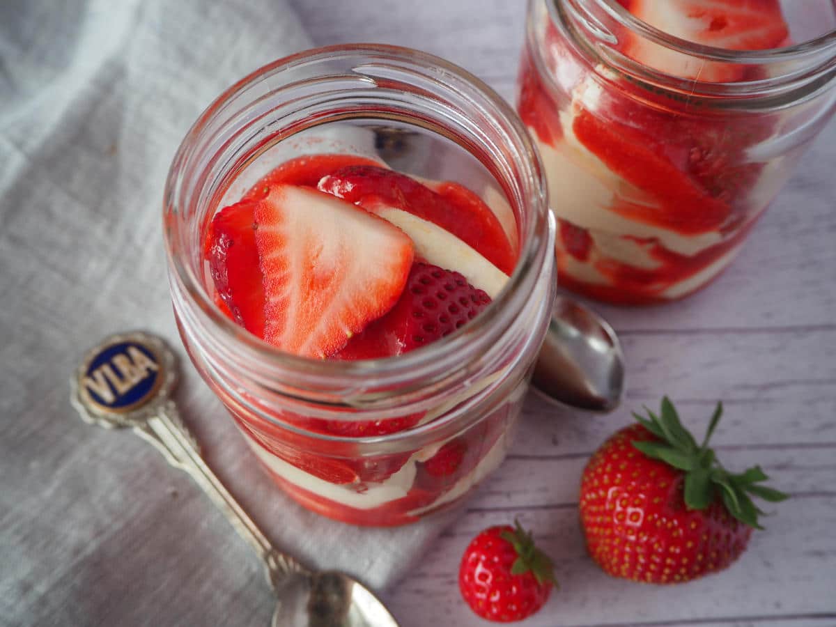 Two jars of Eton mess with fresh strawberries on the side, spoons and a linen cloth.