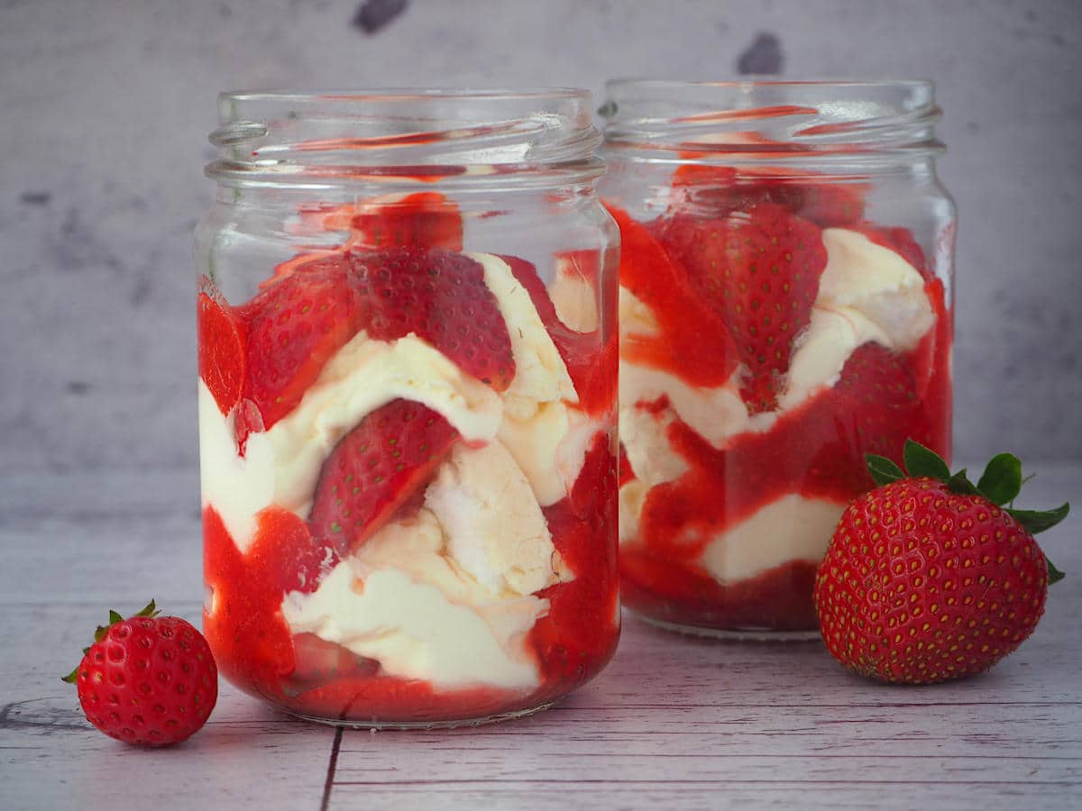 Two jars of Eton mess with fresh strawberries on the side.