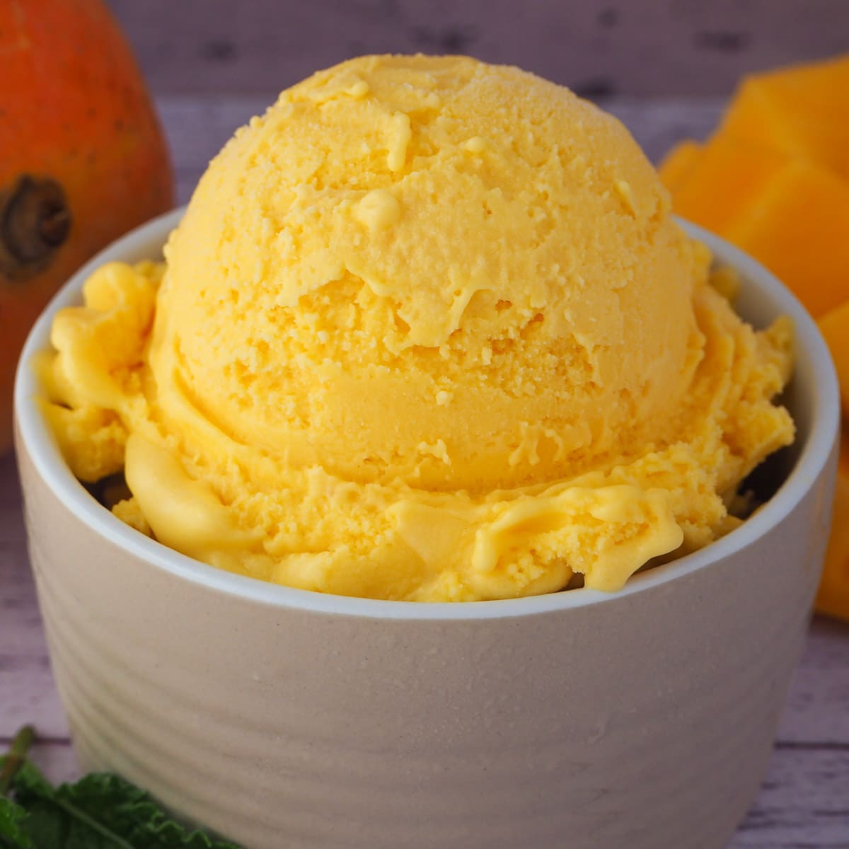 Close up scoop of mango ice cream in a small bow, with fresh mango in the background.